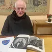 photo  le père jacques-marie guilmard devant le blason de l’abbaye et des portraits de dom guéranger, jeune homme puis moine âgé. 
